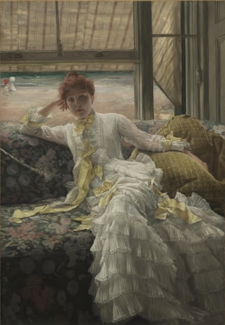 A woman in a luxurious white dress with yellow ribbons lounges on a floral-patterned sofa. She gazes directly forward, resting her head on her hand. The background features a window view of a sandy beach with a couple of figures in the distance.