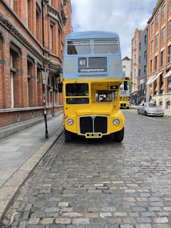 A vintage double-decker bus painted in bright yellow and blue stands on a cobblestone street surrounded by historic red brick buildings. The bus displays advertisements for a vintage tea trip on its front. There is another similar bus further down the street.