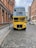 A vintage double-decker bus painted in bright yellow and blue stands on a cobblestone street surrounded by historic red brick buildings. The bus displays advertisements for a vintage tea trip on its front. There is another similar bus further down the street.