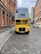 A vintage double-decker bus painted in bright yellow and blue stands on a cobblestone street surrounded by historic red brick buildings. The bus displays advertisements for a vintage tea trip on its front. There is another similar bus further down the street.