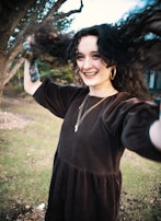 A person with long curly hair is joyfully moving their hair, smiling broadly. They are standing outdoors with a background of trees and grass. The individual is wearing a dark velvet dress, gold hoop earrings, and layered necklaces. A wrist tattoo is visible, and their movement is energetic and lively.