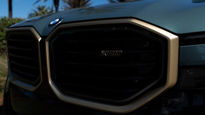The image features a close-up of a car grille with a distinctive design, showcasing an emblem. The surrounding area has a glossy metallic finish with a gold outline. Trees and sky are faintly visible in the reflections.