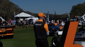 A person wearing McLaren team attire stands in a grassy area with parked race cars nearby. In the background, a crowd mingles near tents and flags, suggesting an outdoor event. The sky is clear and sunny.