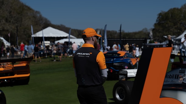 A person wearing McLaren team attire stands in a grassy area with parked race cars nearby. In the background, a crowd mingles near tents and flags, suggesting an outdoor event. The sky is clear and sunny.
