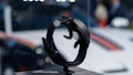 A close-up view of a sleek black smartwatch displayed on a stand in a well-lit environment. The background is blurred, with hints of a vehicle and people, creating a bokeh effect.