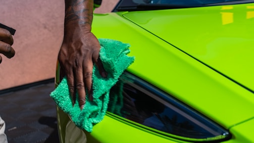 A person is polishing a bright green car with a turquoise cloth. Their hand is visible, showing a tattoo, and they appear to be focused on cleaning the car's headlight area.
