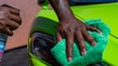 A close-up of a person's hand is polishing a bright green car using a green cloth. Another hand holds a bottle of car cleaning spray. The focus is on the action of cleaning the car's exterior surface near the headlight.