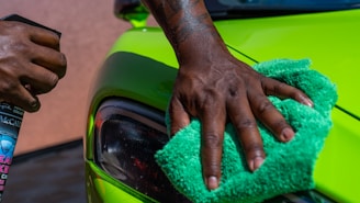 A close-up of a person's hand is polishing a bright green car using a green cloth. Another hand holds a bottle of car cleaning spray. The focus is on the action of cleaning the car's exterior surface near the headlight.