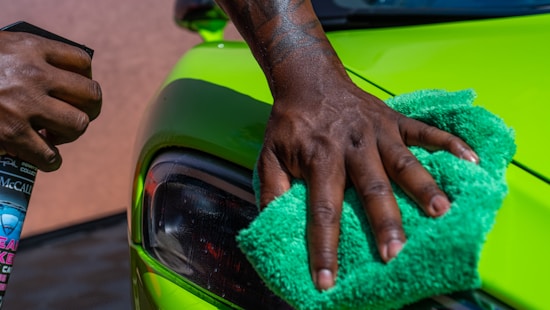 A close-up of a person's hand is polishing a bright green car using a green cloth. Another hand holds a bottle of car cleaning spray. The focus is on the action of cleaning the car's exterior surface near the headlight.