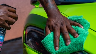 A close-up of a person's hand is polishing a bright green car using a green cloth. Another hand holds a bottle of car cleaning spray. The focus is on the action of cleaning the car's exterior surface near the headlight.