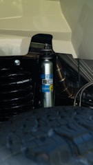 A close-up view of a Bilstein shock absorber mounted on a vehicle, revealing the company logo and various mechanical components. The tire tread is visible in the foreground, indicating this is part of a vehicle's suspension system.