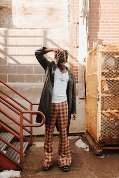 A person dressed in a stylish outfit with a leather coat, plaid pants, and a white shirt holds a vintage camera up to their face. They are standing in an industrial-looking setting with brick and concrete walls, red metal stairs, and a yellow cage-like structure labeled 'empty cylinders'. Shadows and sunlight create an intriguing play of light.