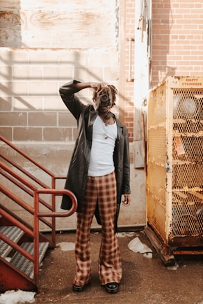A person dressed in a stylish outfit with a leather coat, plaid pants, and a white shirt holds a vintage camera up to their face. They are standing in an industrial-looking setting with brick and concrete walls, red metal stairs, and a yellow cage-like structure labeled 'empty cylinders'. Shadows and sunlight create an intriguing play of light.