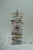 A diverse and colorful collection of wooden signs mounted vertically, each displaying different place names and distances. Set against a misty, mountainous background, the signs point in various directions, creating a striking contrast with the snowy terrain.