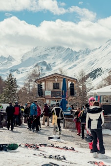 A snowy mountain resort scene featuring a group of people gathered near a wooden chalet. The sky is partly cloudy and the mountains in the background are covered in fresh snow. Ski equipment lies on the ground while individuals, dressed in winter gear, socialize and prepare for skiing.