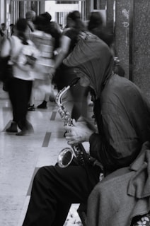 A street musician wearing a hood and sitting on a bench plays a saxophone in a bustling corridor. The background is filled with blurry, motion-captured pedestrians walking by, suggesting a busy environment. The image is monochromatic, adding a somber and artistic feel.