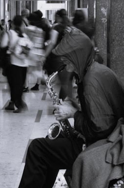 A street musician wearing a hood and sitting on a bench plays a saxophone in a bustling corridor. The background is filled with blurry, motion-captured pedestrians walking by, suggesting a busy environment. The image is monochromatic, adding a somber and artistic feel.