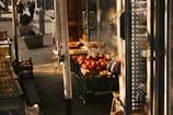 A market stall displays a variety of fresh produce, including tomatoes and red bell peppers, stacked in crates. The scene is warmly lit by afternoon sunlight, casting shadows. In the background, two people walk down the street alongside large planters with small shrubs. Glass-fronted refrigerators and a Coca-Cola advertisement are also visible.