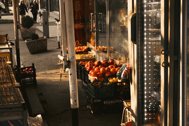 A market stall displays a variety of fresh produce, including tomatoes and red bell peppers, stacked in crates. The scene is warmly lit by afternoon sunlight, casting shadows. In the background, two people walk down the street alongside large planters with small shrubs. Glass-fronted refrigerators and a Coca-Cola advertisement are also visible.