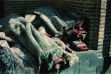 A stack of intricately woven rugs lies against a brick wall. The rugs are partially unrolled, revealing detailed patterns with flourishes and motifs in shades of blue, red, and beige. The fabric is textured, suggesting a traditional or vintage style, and the light casts soft shadows across their surface.