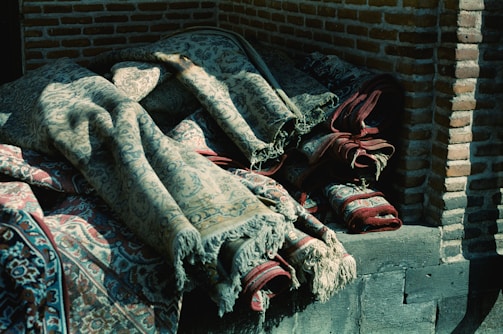 A stack of intricately woven rugs lies against a brick wall. The rugs are partially unrolled, revealing detailed patterns with flourishes and motifs in shades of blue, red, and beige. The fabric is textured, suggesting a traditional or vintage style, and the light casts soft shadows across their surface.