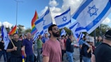 A group of people are participating in a public gathering, holding Israeli flags and rainbow flags. The scene is outdoors with a clear sky and some clouds visible. The crowd appears diverse and engaged in the event, with expressions of solidarity and presence.
