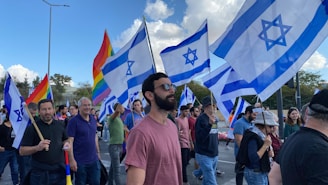 A group of people are participating in a public gathering, holding Israeli flags and rainbow flags. The scene is outdoors with a clear sky and some clouds visible. The crowd appears diverse and engaged in the event, with expressions of solidarity and presence.