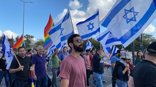 A group of people are participating in a public gathering, holding Israeli flags and rainbow flags. The scene is outdoors with a clear sky and some clouds visible. The crowd appears diverse and engaged in the event, with expressions of solidarity and presence.
