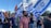 A group of people are participating in a public gathering, holding Israeli flags and rainbow flags. The scene is outdoors with a clear sky and some clouds visible. The crowd appears diverse and engaged in the event, with expressions of solidarity and presence.