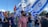 A group of people are participating in a public gathering, holding Israeli flags and rainbow flags. The scene is outdoors with a clear sky and some clouds visible. The crowd appears diverse and engaged in the event, with expressions of solidarity and presence.