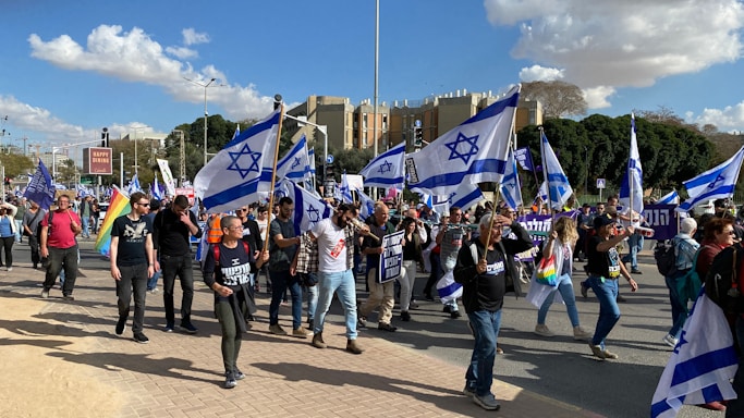 A group of people march holding Israeli flags and banners. Participants wear casual clothing, some wearing shirts with slogans. The setting is an urban area with buildings and trees in the background. The sky is partly cloudy.
