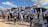A group of people march holding Israeli flags and banners. Participants wear casual clothing, some wearing shirts with slogans. The setting is an urban area with buildings and trees in the background. The sky is partly cloudy.