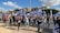 A group of people march holding Israeli flags and banners. Participants wear casual clothing, some wearing shirts with slogans. The setting is an urban area with buildings and trees in the background. The sky is partly cloudy.