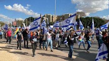 A group of people march holding Israeli flags and banners. Participants wear casual clothing, some wearing shirts with slogans. The setting is an urban area with buildings and trees in the background. The sky is partly cloudy.