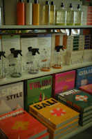 Various bottles of cleaning products are neatly organized on shelves, accompanied by colorful books with travel-themed covers. The upper shelf features sleek bottles with liquids in different hues, while the lower shelf displays vibrant books with titles such as 'Tulum', 'Bali', and 'Jaipur'. The arrangement suggests a boutique or specialty store layout.