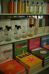 Various bottles of cleaning products are neatly organized on shelves, accompanied by colorful books with travel-themed covers. The upper shelf features sleek bottles with liquids in different hues, while the lower shelf displays vibrant books with titles such as 'Tulum', 'Bali', and 'Jaipur'. The arrangement suggests a boutique or specialty store layout.