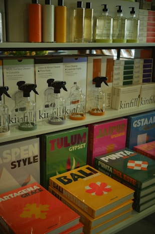 Various bottles of cleaning products are neatly organized on shelves, accompanied by colorful books with travel-themed covers. The upper shelf features sleek bottles with liquids in different hues, while the lower shelf displays vibrant books with titles such as 'Tulum', 'Bali', and 'Jaipur'. The arrangement suggests a boutique or specialty store layout.