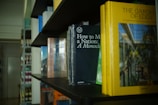A bookshelf is filled with various books, with the focus on a few that include 'How to Make a Nation: A Monocle' with a dark cover and 'The Garden of Eden' with a yellow cover. The setting appears to be a well-lit room, possibly a library or bookstore.