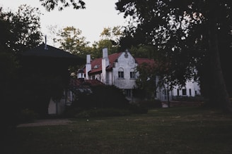 A large, old house with a red tiled roof is surrounded by trees and greenery. The building features multiple chimneys and arched windows, and it appears to be nestled within a wooded area, creating a serene and secluded atmosphere.