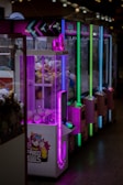 A row of brightly lit arcade claw machines is seen, each with colorful neon lights in purple, green, and blue. The machines are filled with plush toys and other prizes. The front machine has an anime-style design with a character and text that reads 'Fried Eggs.' The environment appears to be dimly lit, highlighting the colorful lights from the machines.