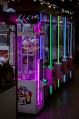 A row of brightly lit arcade claw machines is seen, each with colorful neon lights in purple, green, and blue. The machines are filled with plush toys and other prizes. The front machine has an anime-style design with a character and text that reads 'Fried Eggs.' The environment appears to be dimly lit, highlighting the colorful lights from the machines.