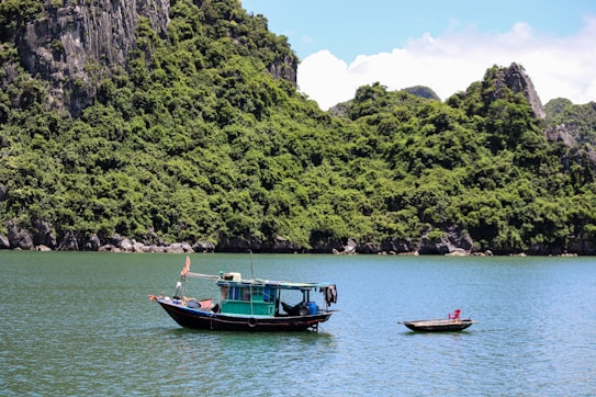 A traditional wooden boat with a colorful cabin floats on calm, turquoise waters. A lush, green, mountainous landscape serves as a backdrop, showcasing dense foliage on rocky hillsides under a bright blue sky.