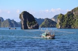 A fishing boat sails on a calm sea, surrounded by towering limestone islands covered in lush green vegetation under a clear blue sky.