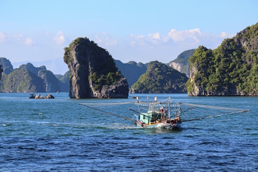 A fishing boat sails on a calm sea, surrounded by towering limestone islands covered in lush green vegetation under a clear blue sky.