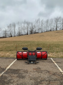 A red snow plow attachment is positioned on an asphalt parking lot, with a backdrop of leafless trees and a grassy hill under a cloudy sky.