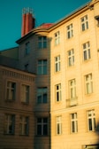 A multi-story residential building with classic architecture is bathed in warm sunlight. The structure features numerous windows and a red brick chimney that stands out against the turquoise sky. The facade has a beige color, and the light casts distinct shadows and highlights, indicating the time of day to be either early morning or late afternoon.