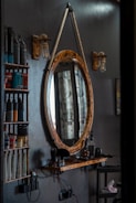 A barbershop setting featuring a rustic, oval-shaped mirror with a thick rope used as a hanger. Adjacent are two wooden shelves holding various hair products, including bottles and tubes. Below the mirror, a wooden ledge supports hair grooming tools like clippers. The scene is dimly lit by vintage-style light bulbs attached to wooden mounts.