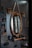 A barbershop setting featuring a rustic, oval-shaped mirror with a thick rope used as a hanger. Adjacent are two wooden shelves holding various hair products, including bottles and tubes. Below the mirror, a wooden ledge supports hair grooming tools like clippers. The scene is dimly lit by vintage-style light bulbs attached to wooden mounts.