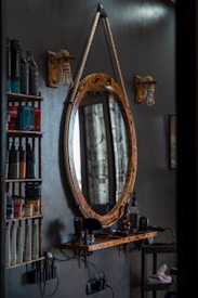 A barbershop setting featuring a rustic, oval-shaped mirror with a thick rope used as a hanger. Adjacent are two wooden shelves holding various hair products, including bottles and tubes. Below the mirror, a wooden ledge supports hair grooming tools like clippers. The scene is dimly lit by vintage-style light bulbs attached to wooden mounts.