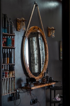 A barbershop setting featuring a rustic, oval-shaped mirror with a thick rope used as a hanger. Adjacent are two wooden shelves holding various hair products, including bottles and tubes. Below the mirror, a wooden ledge supports hair grooming tools like clippers. The scene is dimly lit by vintage-style light bulbs attached to wooden mounts.